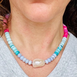 May include: A beaded necklace featuring a large, off-white, irregular-shaped pearl pendant. The necklace has a gradient of blue, turquoise, and pink beads. The necklace is worn around the neck.