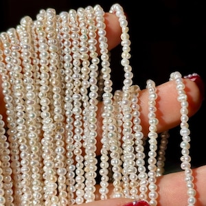 Może przedstawiać: Pasemka małych, kremowobiałych pereł. Perły są nawleczone, tworząc wiele linii delikatnej biżuterii. Perły są trzymane na ciemnym tle, podkreślając ich kolor i fakturę. Obraz jest dobrze oświetlony, ukazując połysk pereł.