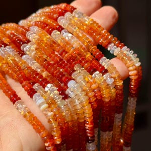 May include: Close-up of multiple strands of gemstone beads. The beads are small and round, in shades of orange, red, and clear. The colours vary from deep red to pale orange and clear, creating a gradient effect. The beads are strung together to create necklaces or bracelets.