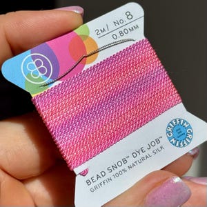 May include: A spool of pink and red silk thread, labeled "BEAD SNOB DYE JOB" and "GRIFFIN 100% NATURAL SILK". The label also indicates the thread is 2m/No. 8, with a diameter of 0.80mm. The spool has a colorful design.