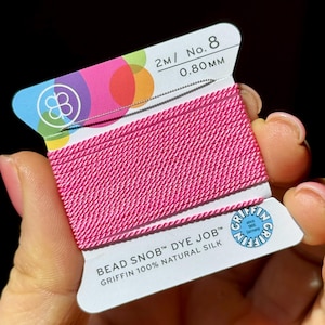 May include: A spool of bright pink Griffin Bead Snob dye job silk thread. The label reads "2M/No. 8 0.80mm" and "Griffin 100% Natural Silk". The spool is held in a hand.