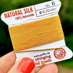 Könnte beinhalten: Eine Spule mit Naturseidenfaden, 100% Naturseide, in leuchtend gelber Farbe. Die Spule ist mit "No. 6 Ø 0,70 mm" und "www.griffin.de stringing and beads" beschriftet.
