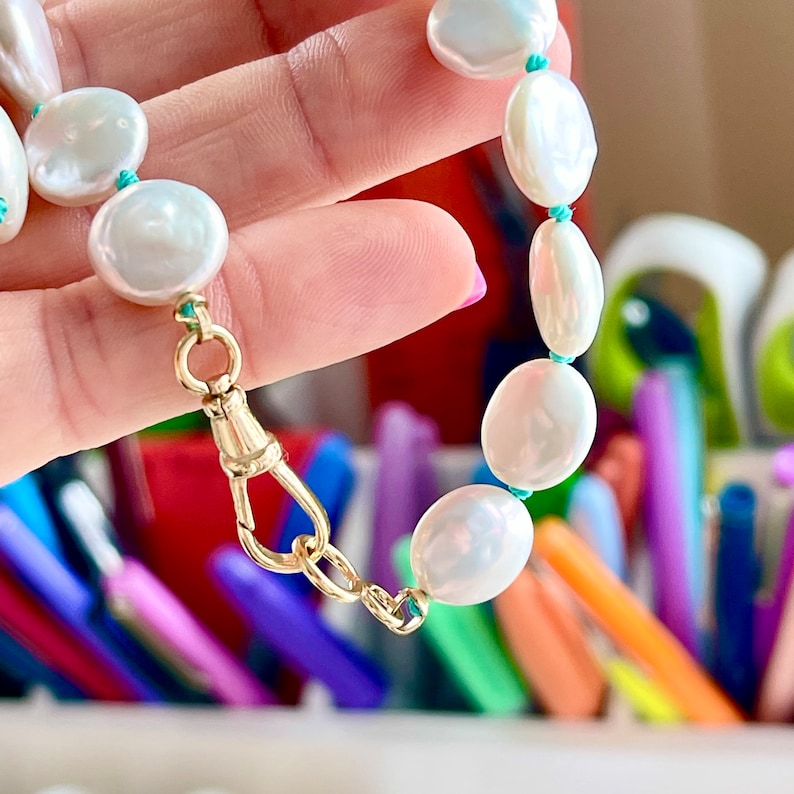 May include: A white pearl necklace with a gold clasp and turquoise string. The necklace is being held by a hand.
