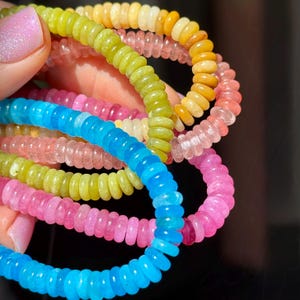 May include: A set of four colorful beaded bracelets, each with a different color: blue, pink, green, and yellow. The bracelets are made of small, round beads that are strung together in a spiral pattern.