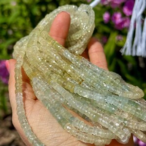 Pu&ograve; includere: Fili di perline di pietre preziose verde chiaro e giallo. Le perline sono piccole, tagliate a forma quadrata e infilate insieme. Le perline sono tenute in una mano, con un filo bianco e uno sfondo sfocato di fiori rosa.