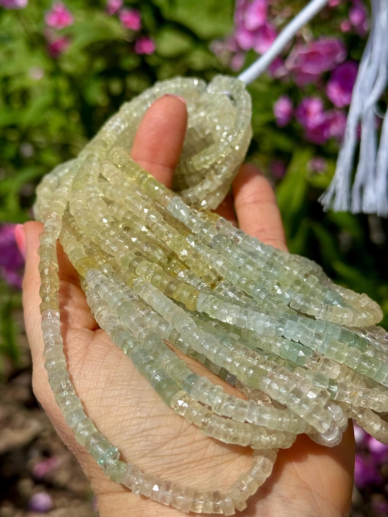 Pu&ograve; includere: Fili di perline di pietre preziose verde chiaro e giallo. Le perline sono piccole, tagliate a forma quadrata e infilate insieme. Le perline sono tenute in una mano, con un filo bianco e uno sfondo sfocato di fiori rosa.