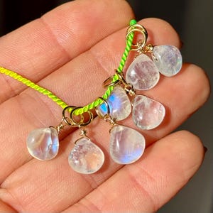 Op de afbeelding: Een handgemaakte ketting met druppelvormige maansteen hangers. De hangers zijn goudkleurig bedraad en geregen op een neon groen koord. De maanstenen hebben een doorschijnende, iriserende kwaliteit.