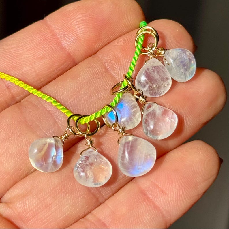 Op de afbeelding: Een handgemaakte ketting met druppelvormige maansteen hangers. De hangers zijn goudkleurig bedraad en geregen op een neon groen koord. De maanstenen hebben een doorschijnende, iriserende kwaliteit.