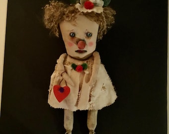 May include: A handmade doll with a painted face, curly hair, and a white dress. The doll has a red nose and lips, and a heart-shaped purse. A flower and leaves adorn the doll's head. The doll is a folk art creation.