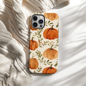 May include: A phone case with a repeating pattern of orange pumpkins and green leaves on a cream background. The pumpkins have green stems. The case is on a cream-coloured knitted blanket.