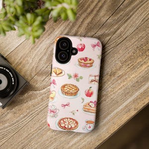 K&ouml;nnte beinhalten: Eine rosa Handyh&uuml;lle mit einem Muster aus Torten, &Auml;pfeln und Schleifen. Die H&uuml;lle hat einen Kameraausschnitt und einen wei&szlig;en Hintergrund.