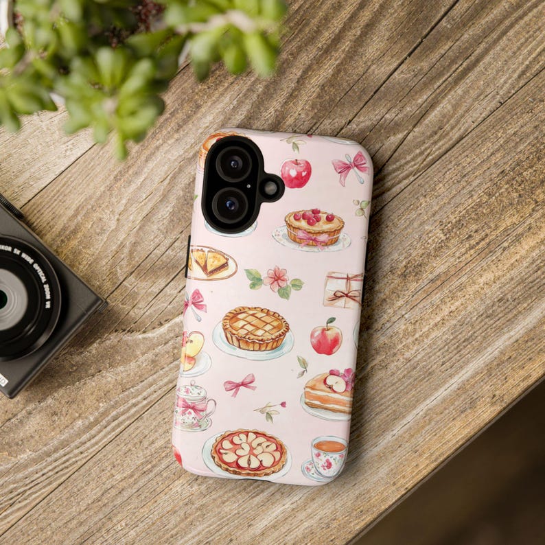 K&ouml;nnte beinhalten: Eine rosa Handyh&uuml;lle mit einem Muster aus Torten, &Auml;pfeln und Schleifen. Die H&uuml;lle hat einen Kameraausschnitt und einen wei&szlig;en Hintergrund.