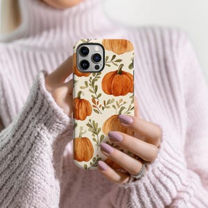 May include: A phone case with a repeating pattern of orange pumpkins and green leaves on a cream background. The case is held in a person's hands, showcasing the autumn design. The case has a glossy finish.