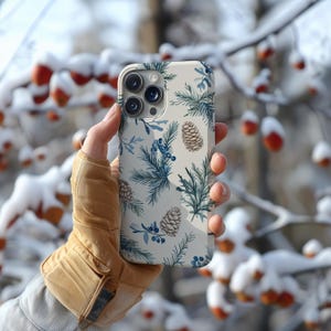 May include: A white phone case with a wintery design featuring pine branches, pine cones, and blue berries.