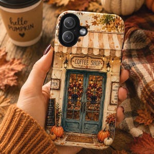 Op de afbeelding: Een telefoonhoesje met een aquarelillustratie van een koffiewinkelgevel met een gestreepte luifel, dubbele deuren versierd met herfstkransen, pompoenen op de grond en borden met de tekst COFFEE SHOP, OPEN en WELCOME.