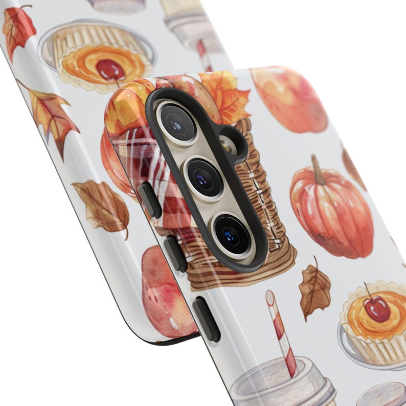 May include: A white phone case with a fall-themed design. The case features a basket of apples, pumpkins, leaves, and a cup of coffee. The phone case has a cutout for the camera lens.