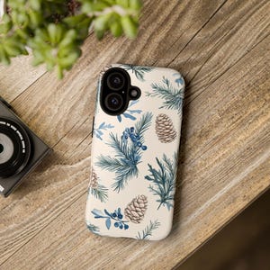 May include: A white phone case with a wintery design featuring blue pine branches, brown pine cones, and small blue berries.