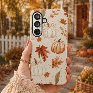 May include: A cream-colored smartphone case featuring a repeating pattern of white and orange pumpkins, brown and orange autumn leaves, and small gold glitter accents. The case has a triple-lens camera cutout on the upper left side.
