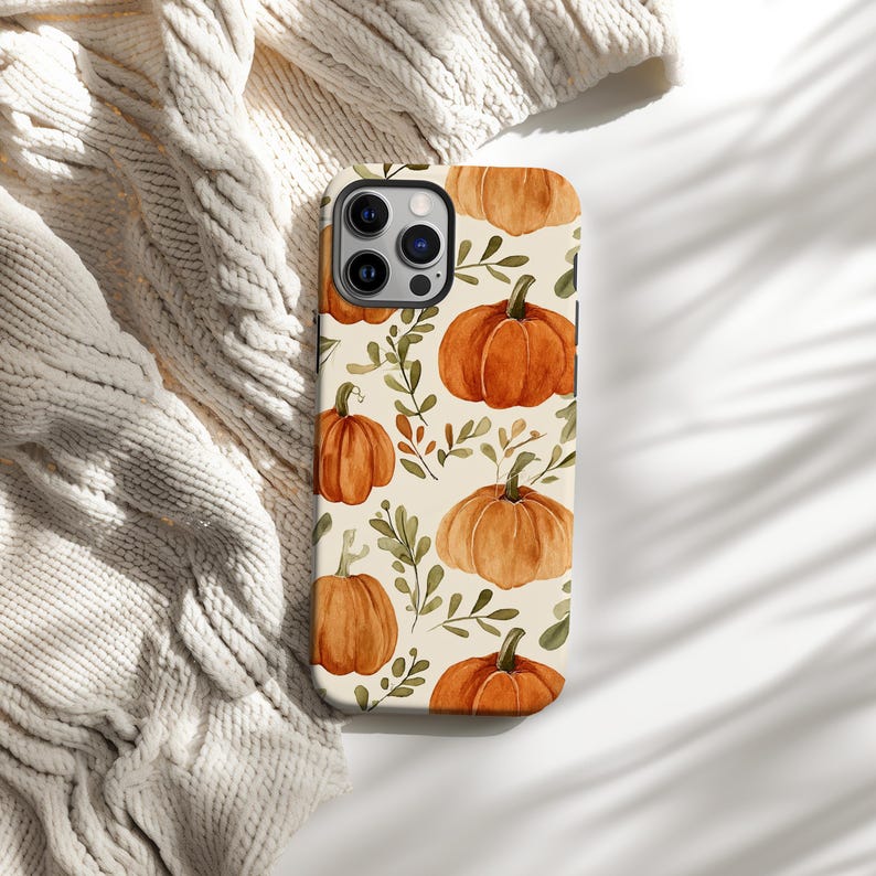 May include: A phone case with a repeating pattern of orange pumpkins and green leaves on a cream background. The pumpkins have green stems. The case is on a cream-coloured knitted blanket.