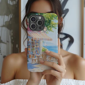May include: A colorful phone case with a watercolor painting of a bookstore with a striped awning, a sailboat in the distance, and a blue sky. The case has a cutout for the phone's camera.