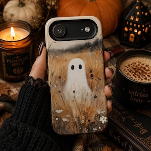 May include: A rectangular phone case featuring a printed illustration of a white ghost standing in a field of dried grasses and small white flowers. The background of the print is a warm, muted golden-brown tone with a dark horizon line at the top.