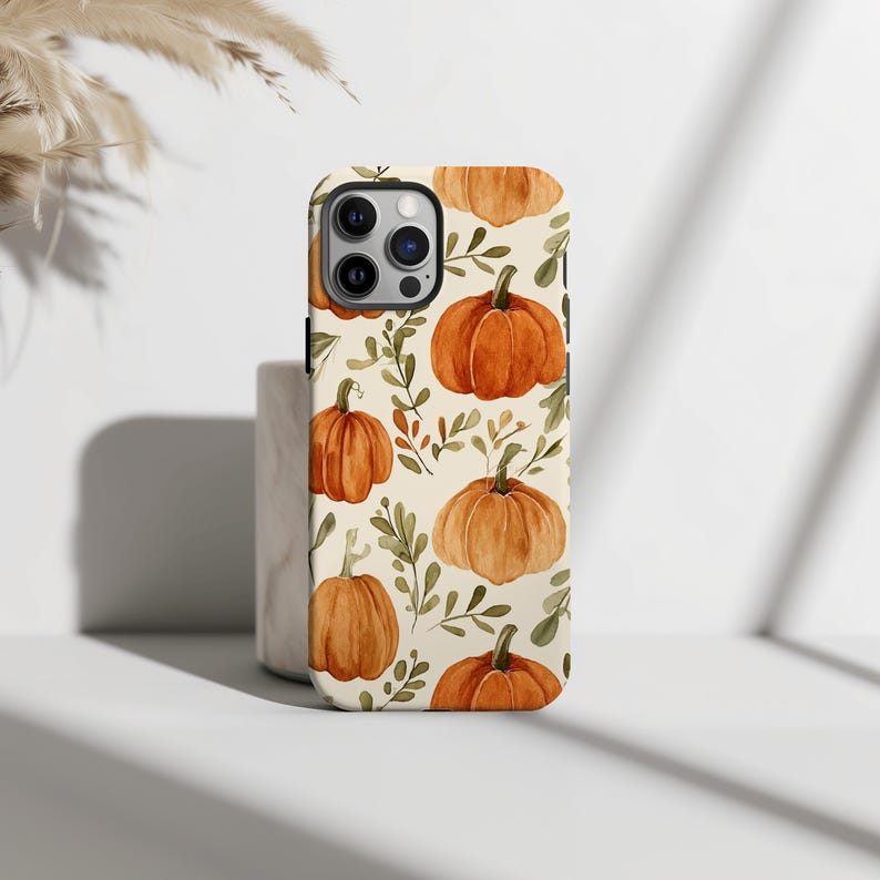 May include: A phone case with a repeating pattern of orange pumpkins and green leaves on a cream background. The case has a glossy finish and a camera cutout. The pumpkins are in a watercolor style, creating a seasonal autumn theme.