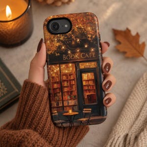 May include: A smartphone case featuring a printed illustration of a cozy book cafe storefront at night. The design displays warm glowing string lights, shelves filled with books, and a small outdoor table with two chairs. The text BOOK CAFE is visible.