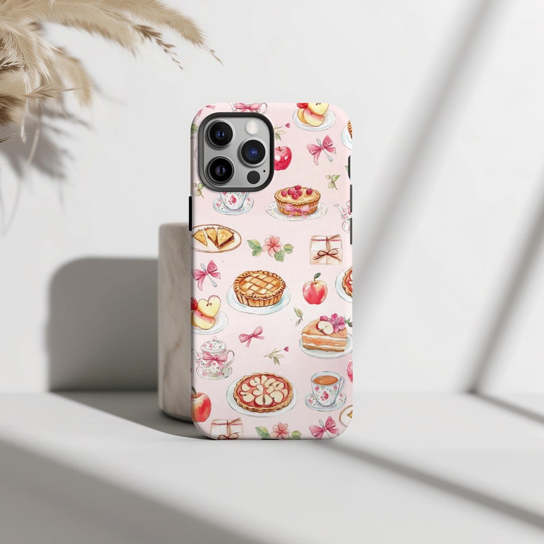 This coquette phone case features charming watercolor illustrations of pies, teacups, apples, and bows on a soft pink background. Its delicate, whimsical design exudes a vintage, feminine charm perfect for adding a touch of elegance.
