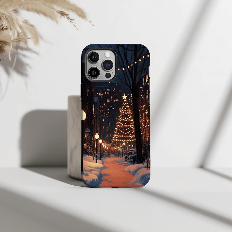 May include: A phone case featuring a winter scene of a snowy street with a lit Christmas tree and fairy lights. The case is black and has a cut-out for the phone's camera.