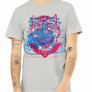 May include: A heather grey t-shirt with a graphic print of a blue and red illustration of a person playing a guitar. The text "Robert Johnson Taught The Devil How To Play Guitar" is printed on the shirt.
