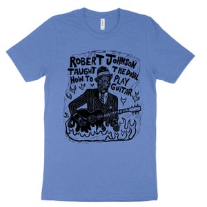 Könnte beinhalten: Ein blaues T-Shirt mit einer schwarz-weißen Illustration von Robert Johnson, der Gitarre spielt, mit dem Text "Robert Johnson Taught The Devil How To Play Guitar".