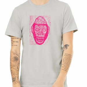 Peut inclure: Un t-shirt gris clair avec un imprimé graphique rose d'un singe avec un cerveau pour tête. Le graphique est dans un style gravure sur bois.