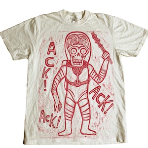 May include: Off-white t-shirt featuring a red graphic of a skeletal alien in a spacesuit holding a staff. The word "ACK!" is printed in red on the left and right sides of the alien. The shirt has short sleeves and a crew neck.