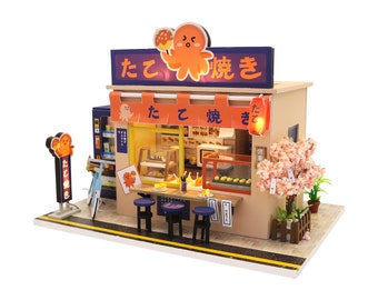 May include: Miniature Japanese takoyaki shop model with a tan exterior, orange awning, and blue accents. The shop features a sign with Japanese text and an octopus mascot. Includes a vending machine, cherry blossom tree, and small stools.