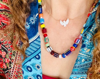 May include: A colorful beaded necklace with rectangular and round beads in various colors, including red, blue, yellow, and green. A gold chain necklace with a white dove pendant is also visible. The person is wearing a red top and a patterned blue and red kimono.