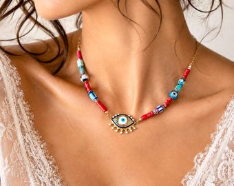 Könnte beinhalten: Eine goldfarbene Halskette mit einem zentralen Schutzaugen-Anhänger mit blau-weiß-schwarzen Emaille-Details und herabhängenden goldenen Strahlen. Die Kette ist mit verschiedenen bunten Perlen verziert, darunter rote Zylinder, gemusterte blaue und türkisfarbene Elemente.