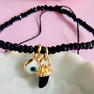 May include: Black beaded bracelet with gold charms. The charms include an eye with a blue iris and a black tooth. The bracelet is displayed on a white, scalloped dish against a pink background. The bracelet is made of small, faceted beads.