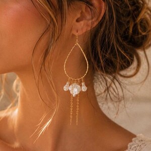 May include: A gold-toned teardrop-shaped earring featuring a textured wire frame. The base of the earring is adorned with three irregular white baroque pearls suspended from delicate gold chains, creating a layered and dangling design.