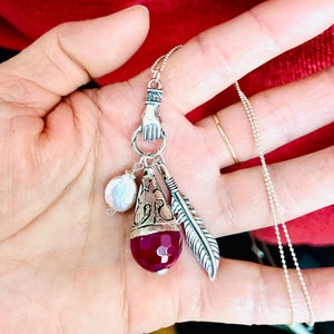 May include: Silver pendant necklace with a hand, feather, faceted red gemstone, and a pearl bead. The necklace has a delicate chain. The background is red.