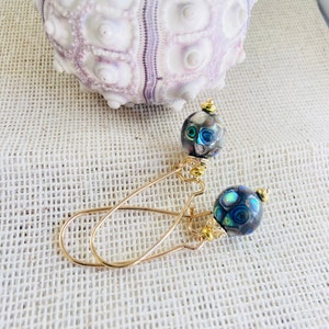 Pode incluir: Brincos pendurados dourados com contas de concha de abalone cinza escuro e iridescente. Os brincos têm um design simples e elegante com fecho de gancho. Uma ouriço-do-mar branco e roxo no fundo.