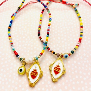 May include: Two colorful beaded bracelets with red string closures. Each bracelet features a gold charm with a red heart and a small evil eye charm.