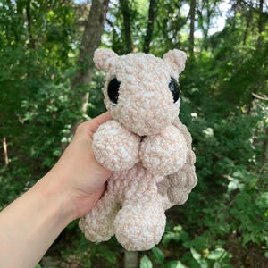 May include: A handmade, plush toy in the shape of a small animal, possibly a squirrel or a similar creature. The toy is primarily light beige with darker beige accents on the back and ears. It has large, black, shiny eyes.
