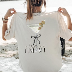 May include: White t-shirt with a martini glass graphic and the text "a bride in a tinibit" printed on the back.