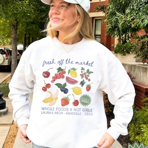 May include: White sweatshirt featuring a fruit-themed graphic with the text "fresh off the market." The design includes illustrations of various fruits. Below the graphic are the words "WHOLE FOODS X HOT GIRLS LAURA'S BACH - ASHEVILLE - 2025."