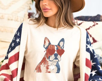 May include: Cream-colored t-shirt featuring a French bulldog wearing heart-shaped sunglasses. The dog's fur is colored in red, white, and blue. The person is wearing a cardigan with an American flag design and denim shorts.