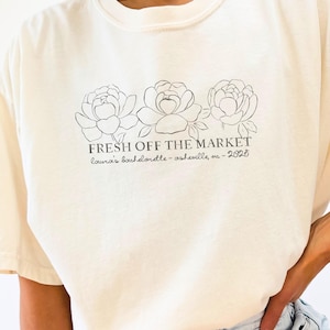 May include: A cream-coloured t-shirt featuring a floral design and the text "FRESH OFF THE MARKET." Below the text is "Laura's bachelorette - asheville, nc - 2025" in a script font. The shirt is worn with denim shorts.