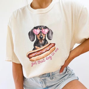 May include: Cream-colored t-shirt featuring a dachshund wearing pink heart-shaped sunglasses and a hot dog. The text "got that dog in me" is printed below the hot dog in pink script. The shirt is a casual style.