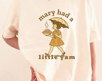 May include: Cream-colored t-shirt featuring a vintage-style illustration of a girl holding a pie under an umbrella. The text "mary had a little yam" is arched above and below the illustration. The shirt has short sleeves.