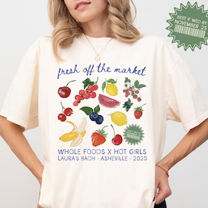 May include: A cream-coloured t-shirt featuring a fruit graphic with cherries, blueberries, lemons, strawberries, and a banana. Text reads "fresh off the market," "WHOLE FOODS X HOT GIRLS LAURA'S BACH ASHEVILLE 2025," and "BEST IF WED BY: NOVEMBER 25."