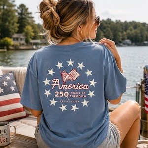 May include: A blue short-sleeve t-shirt featuring a graphic on the back with a circle of white stars surrounding a waving American flag, the text America 250 Years of Freedom, and the dates 1776-2026 printed in white and red ink.